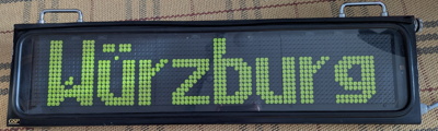 The front side of the display unit. A flip-dot matrix display shows the text "Würzburg" in green text on black background, with some stuck pixels. All in a black metal box with two silver carrying handles.