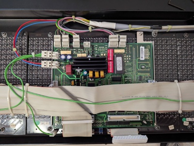 The inside cabling of the display unit, with the control PCB, the back side of the flipdot panels, and cabling. There are several 40-pin ribbon cables stacked on top of each other going from the control PCB to the panels. Some cables are connected to multiple panels. On top, there is a bundle of green wires running to the modules, and a few other cables that lead to the outside connector.